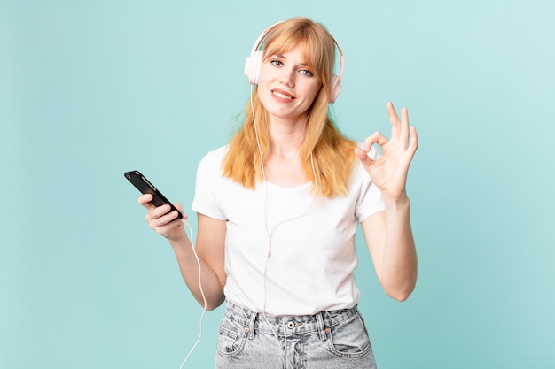 Mujer bonita pelirroja que se siente feliz, mostrando aprobación con un gesto aceptable y escuchando música con auriculares