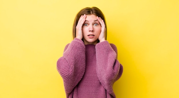 Mujer bonita pelirroja que se siente horrorizada y conmocionada levantando las manos a la cabeza y entrando en pánico por un error