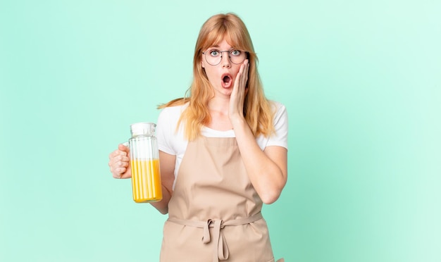 Mujer bonita pelirroja sintiéndose conmocionada y asustada con delantal preparando un jugo de naranja