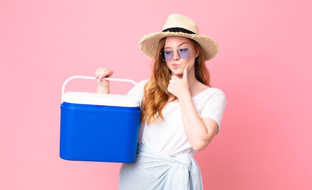 Mujer bonita pelirroja sonriendo con una expresión feliz y segura con la mano en la barbilla y sosteniendo un refrigerador portátil de picnic