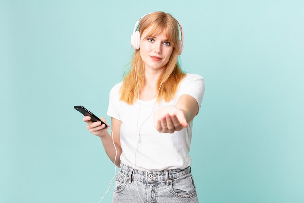 Mujer bonita pelirroja sonriendo felizmente con amigable y ofreciendo y mostrando un concepto y escuchando música con auriculares