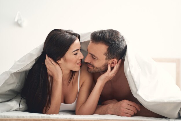 Mujer bonita de pelo oscuro complacida y su marido joven y guapo sonriendo el uno al otro en la cama