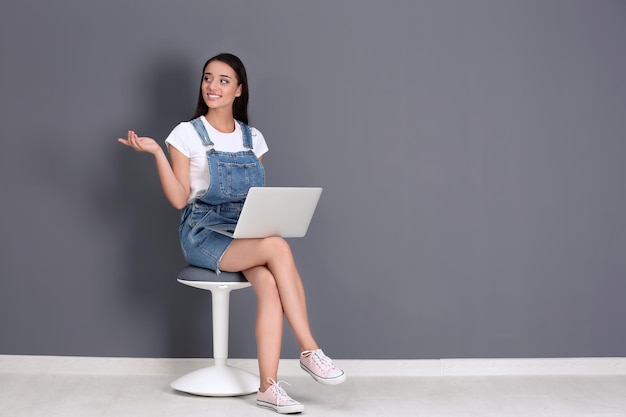 Mujer bonita con portátil moderno sentado en una silla contra la pared gris