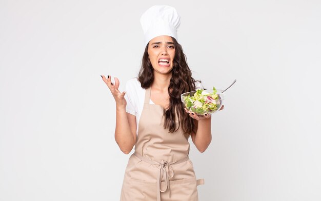 Mujer bonita que parece desesperada, frustrada y estresada usando un delantal y sosteniendo una ensalada