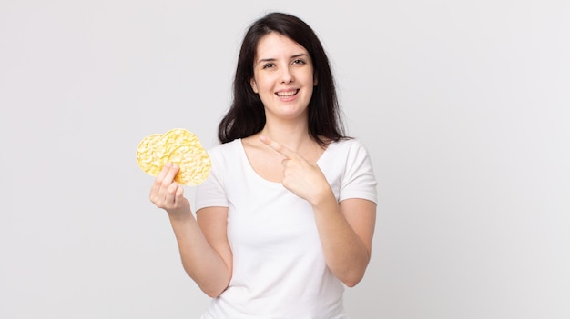 Mujer bonita que parece emocionada y sorprendida apuntando hacia un lado y sosteniendo pasteles de arroz dietéticos