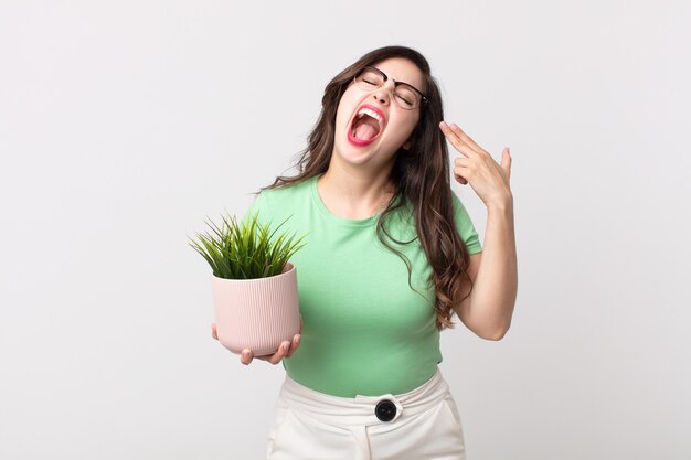 Mujer bonita que parece infeliz y estresada, gesto de suicidio haciendo un signo de pistola y sosteniendo una planta decorativa