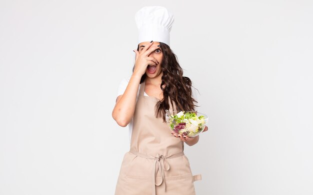 Mujer bonita que parece sorprendida, asustada o aterrorizada, cubriendo la cara con la mano con un delantal y sosteniendo una ensalada