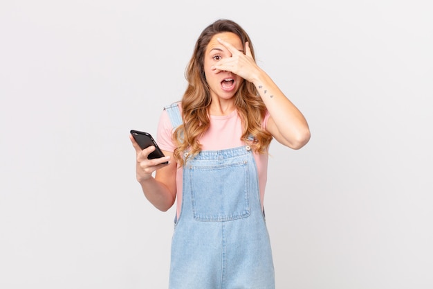 Mujer bonita que parece sorprendida, asustada o aterrorizada, cubriendo la cara con la mano y sosteniendo un teléfono inteligente