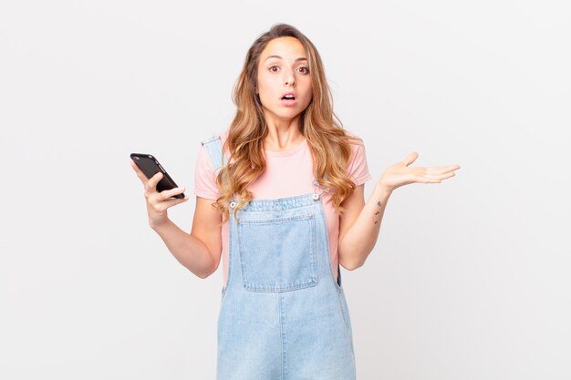 Mujer bonita que parece sorprendida y conmocionada, con la mandíbula caída sosteniendo un objeto y sosteniendo un teléfono inteligente