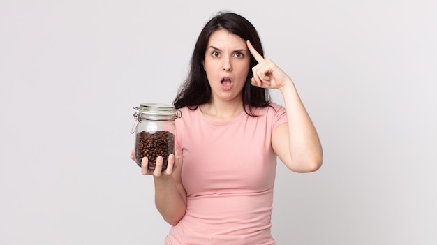 Mujer bonita que parece sorprendida, dándose cuenta de un nuevo pensamiento, idea o concepto y sosteniendo una botella de granos de café