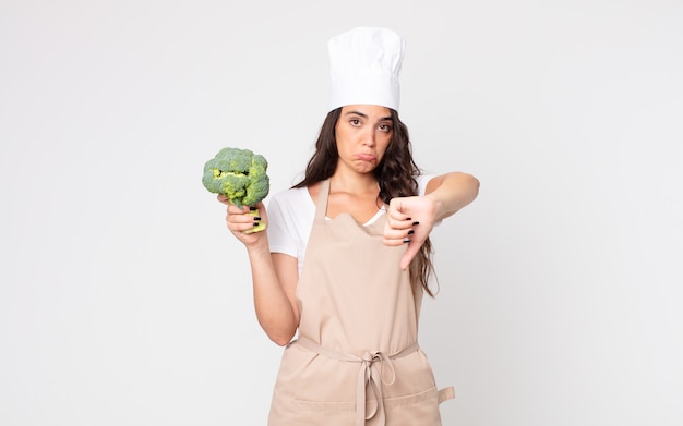 Mujer bonita que se siente cruzada, mostrando los pulgares hacia abajo con un delantal y sosteniendo un brócoli