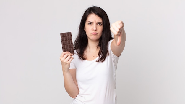 Mujer bonita que se siente cruzada, mostrando los pulgares hacia abajo y sosteniendo una barra de chocolate
