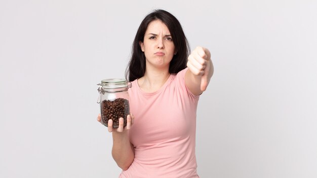 Mujer bonita que se siente cruzada, mostrando los pulgares hacia abajo y sosteniendo una botella de granos de café