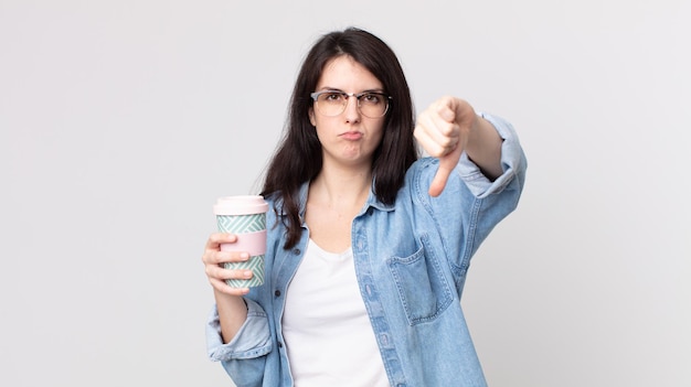 Mujer bonita que se siente cruzada, mostrando los pulgares hacia abajo y sosteniendo un café para llevar