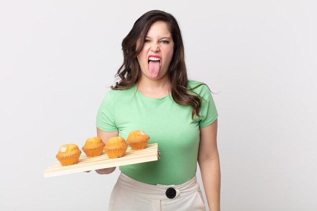 Mujer bonita que se siente disgustada e irritada y con la lengua fuera y sosteniendo una bandeja de muffins