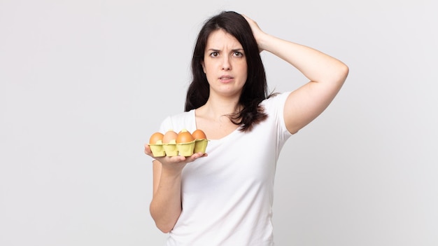 Mujer bonita que se siente estresada, ansiosa o asustada, con las manos en la cabeza y sosteniendo una caja de huevos