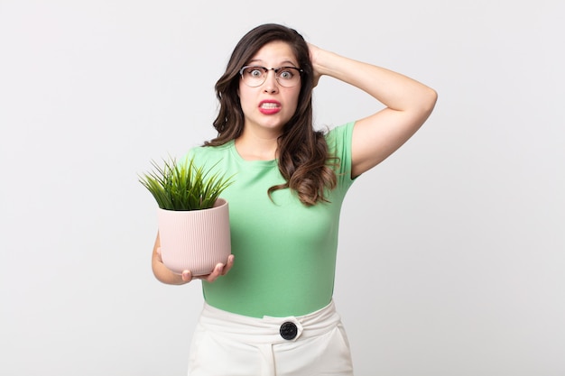 Mujer bonita que se siente estresada, ansiosa o asustada, con las manos en la cabeza y sosteniendo una planta decorativa