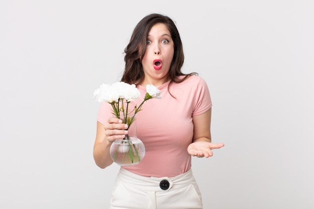 Mujer bonita que se siente extremadamente conmocionada y sorprendida y sosteniendo una maceta de flores decorativas