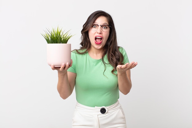 Mujer bonita que se siente extremadamente conmocionada y sorprendida y sosteniendo una planta decorativa