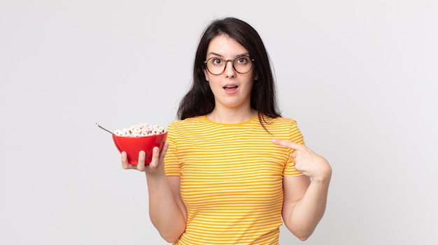 Mujer bonita que se siente feliz y apuntando a sí misma con un emocionado y sosteniendo un tazón de desayuno