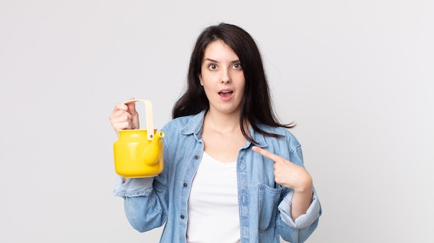 Mujer bonita que se siente feliz y apuntando a sí misma con un emocionado y sosteniendo una tetera