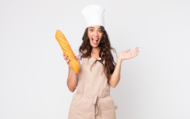 Mujer bonita que se siente feliz y asombrada por algo increíble con un delantal y sosteniendo una barra de pan