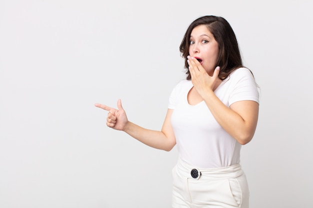 Mujer bonita que se siente feliz, conmocionada y sorprendida, cubriendo la boca con la mano y apuntando al espacio lateral de la copia