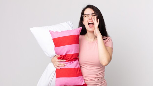 Mujer bonita que se siente feliz, dando un gran grito con las manos al lado de la boca en pijama y sosteniendo una almohada