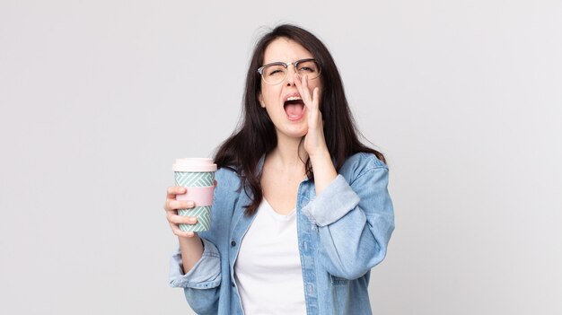 Mujer bonita que se siente feliz, dando un gran grito con las manos al lado de la boca y sosteniendo un café para llevar