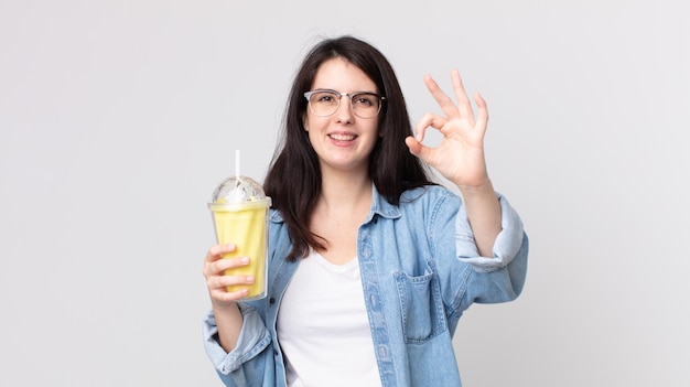 Mujer bonita que se siente feliz, mostrando aprobación con un gesto aceptable y sosteniendo un batido de vainilla