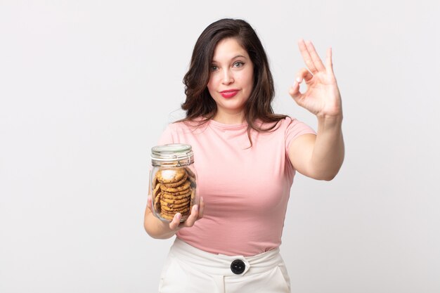 Mujer bonita que se siente feliz, mostrando aprobación con un gesto aceptable y sosteniendo una botella de vidrio de galletas