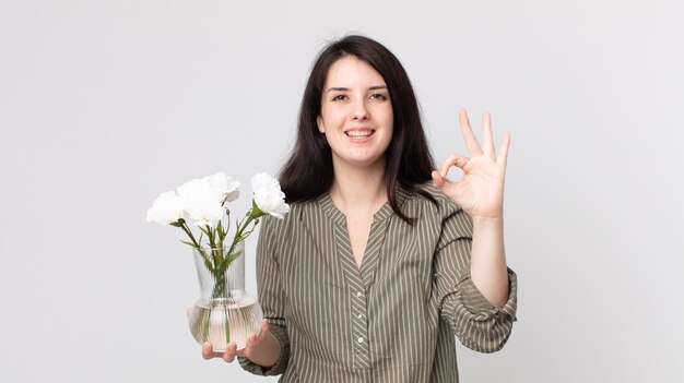 Mujer bonita que se siente feliz, mostrando aprobación con un gesto aceptable y sosteniendo flores decorativas. agente asistente con un auricular