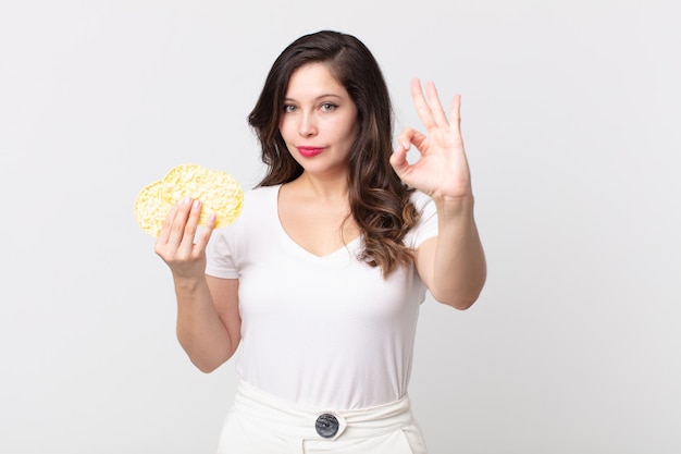 Mujer bonita que se siente feliz mostrando aprobación con un gesto bien y sosteniendo una dieta de pasteles de arroz