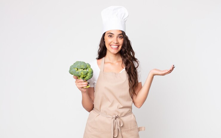 Foto mujer bonita que se siente feliz, sorprendida al darse cuenta de una solución o idea con un delantal y sosteniendo un brócoli