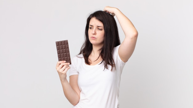 Mujer bonita que se siente perpleja y confundida, rascándose la cabeza y sosteniendo una barra de chocolate