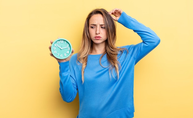 Mujer bonita que se siente perpleja y confundida, rascándose la cabeza sosteniendo un despertador