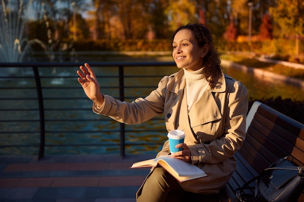 Mujer bonita de raza mixta en traje beige sentada en un banco del parque con una taza de café y un libro de mano, saludando mientras mira a otro lado en el fondo del hermoso lago con hojas de otoño amarillas reflejadas