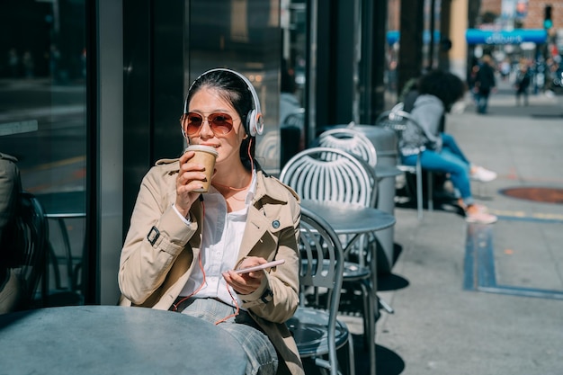 Una mujer bonita se relaja al aire libre en la cafetería de la calle, se sienta en la mesa, escucha música con auriculares y usa el teléfono móvil. feliz mujer despreocupada con gafas de sol bebiendo bebidas de papel bajo el sol en el café