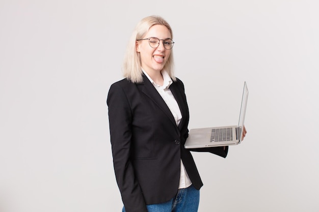 Mujer bonita rubia con actitud alegre y rebelde bromeando y sacando la lengua y sosteniendo una computadora portátil