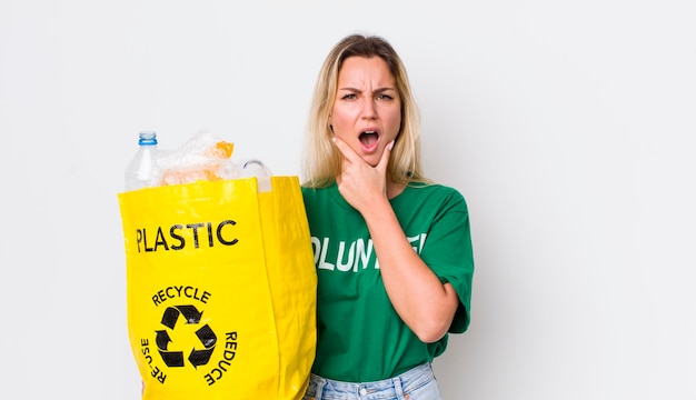 Mujer bonita rubia con la boca y los ojos bien abiertos y la mano en la barbilla concepto de reciclaje