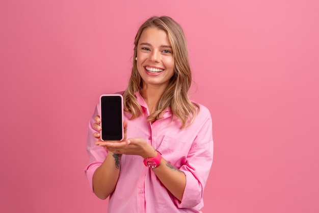 Mujer bonita rubia en camisa rosa sonriendo sosteniendo usando smartphone posando en rosa sonriendo divirtiéndose, mostrando la pantalla del teléfono