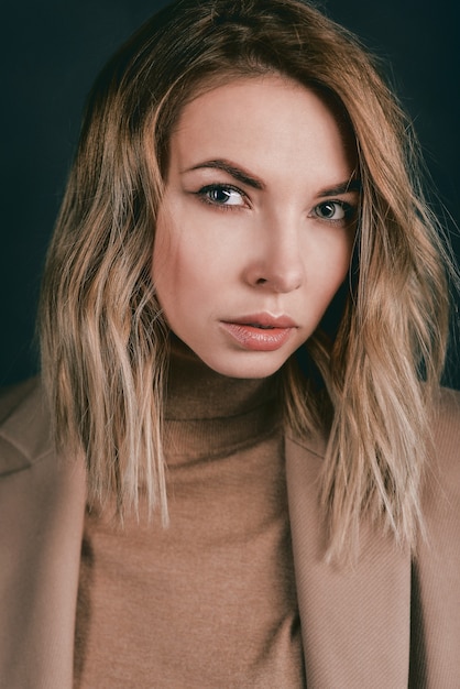 Mujer bonita rubia en la chaqueta beige y camisa de cuero sobre el fondo oscuro