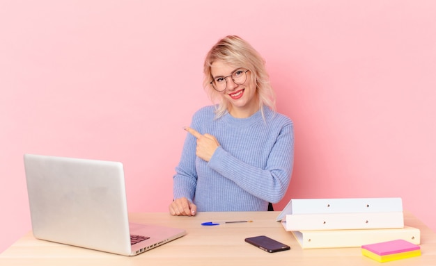 Mujer bonita rubia joven mujer bonita sonriendo alegremente, sintiéndose feliz y apuntando hacia un lado. concepto de escritorio de espacio de trabajo