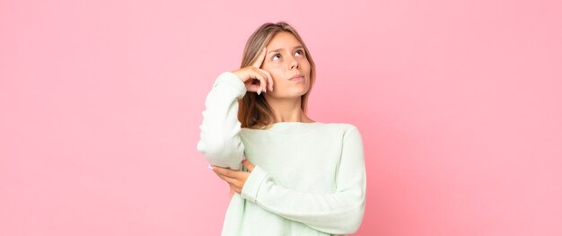 Mujer bonita rubia con una mirada concentrada, preguntándose con una expresión dudosa, mirando hacia arriba y hacia un lado