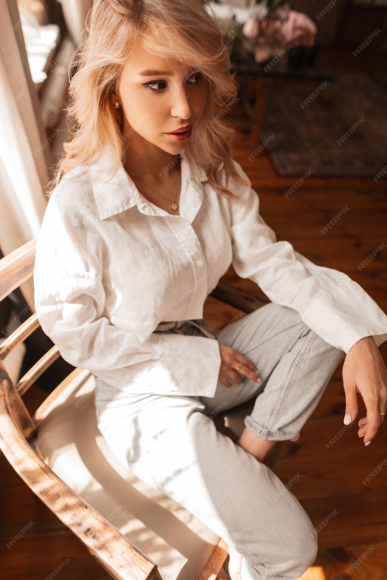 Mujer bonita rubia de moda con camisa blanca y pantalones de mezclilla se sienta una silla de madera en rom con luz solar | Foto Premium