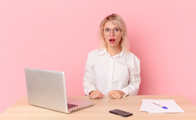 Mujer bonita rubia mujer bonita joven que parece muy conmocionada o sorprendida. concepto de escritorio de espacio de trabajo