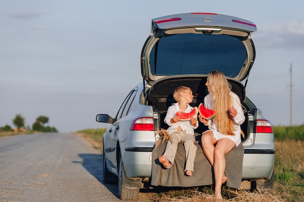 Mujer bonita rubia con pequeño hijo rubio al atardecer relajarse detrás del coche y comer sandía. verano, viajes, naturaleza y aire fresco en el campo.