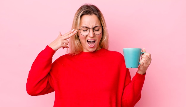 Mujer bonita rubia que parece infeliz y estresada gesto de suicidio haciendo concepto de taza de café de signo de pistola