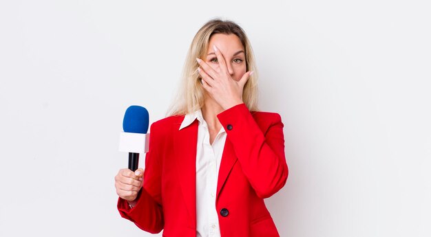 Mujer bonita rubia que parece sorprendida asustada o aterrorizada cubriendo la cara con el preportador de mano y el concepto de micrófono