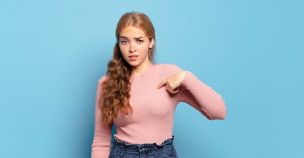 Mujer bonita rubia que se siente confundida, perpleja e insegura, señalando a sí misma preguntándose quién, yo?